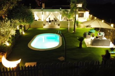 Piscina interrata Skysand Ovale 600 con bordo bianco antiscivolo e ampio solarium in erba artificiale, completata da zona lounge con divani e ombrellone per momenti conviviali all’aperto.