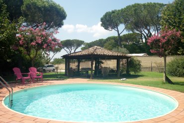 Piscina interrata in lamiera d'acciaio Skysand Tonda 600 con liner color sabbia, bordo effetto terracotta e ampio giardino con pini e fioriture estive. Un angolo di relax immerso nel verde.