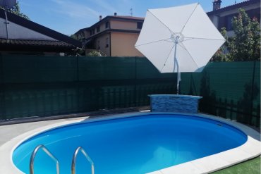 Piscina interrata in lamiera d’acciaio con rivestimento azzurro Skyblue, dotata di elegante parete cascata rivestita in mosaico e ombrellone bianco per un’oasi di relax.
